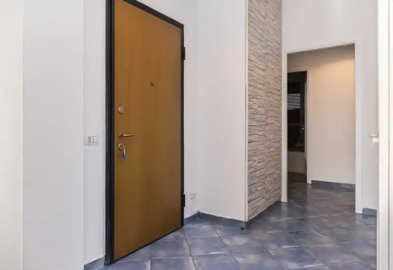 Bright, clean entrance corridor with a wooden front door, tiled floor and a doorway leading to other rooms — practical and well-lit entry area.