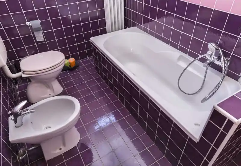 Bright, tiled bathroom featuring a bathtub with hand shower, toilet and bidet on glossy purple tiles.