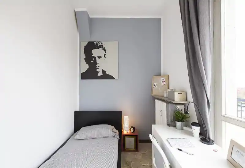 Piccola camera singola arredata con gusto: letto con struttura nera, lampada sul comodino e scrivania vicino alla grande finestra che illumina l'ambiente con luce naturale.
