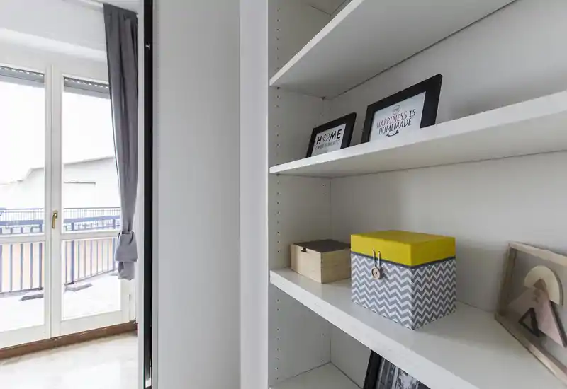Zona giorno luminosa con ampia porta sul balcone e scaffalatura integrata con cornici decorative e scatole contenitive — un angolo moderno e pulito perfetto per esporre e godere della luce.