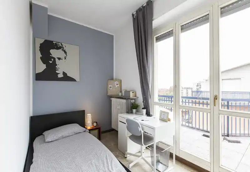 Camera singola luminosa e moderna con letto ben rifatto, lampada sul comodino e una scrivania compatta accanto a grandi finestre con balcone — ideale per uno studente o occupante singolo.