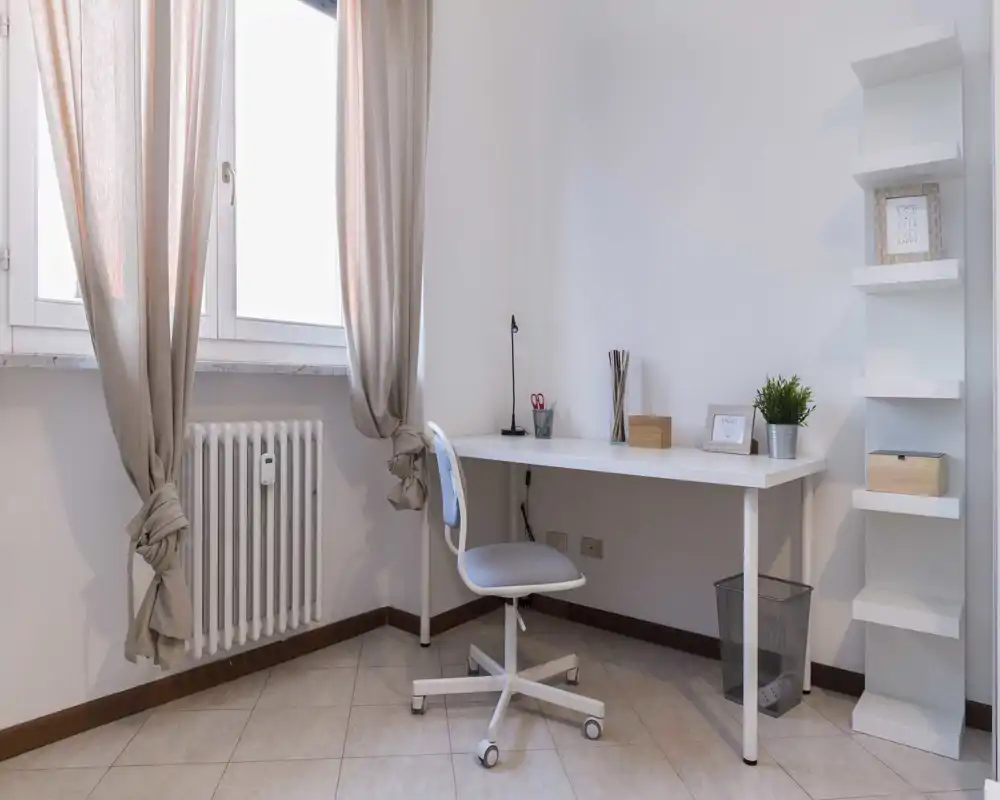 Angolo studio minimale e luminoso con scrivania bianca, sedia girevole, mensole ordinate e luce naturale dalla finestra con tende — perfetto per lavorare concentrati.