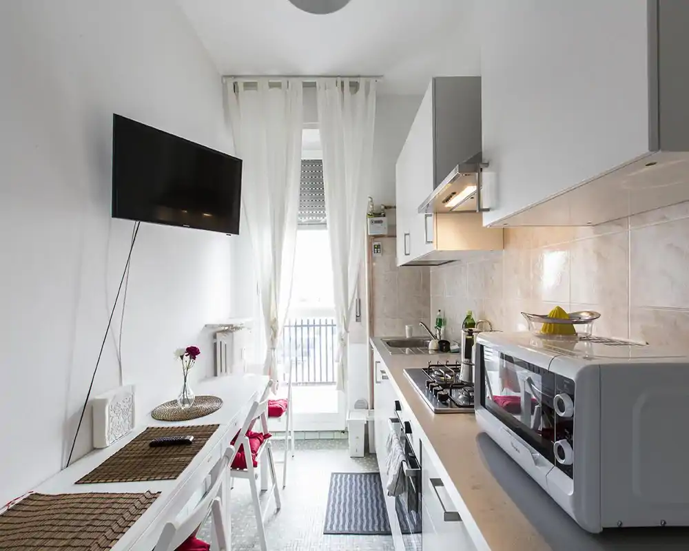 Cucina moderna a corridoio luminosa con mobili bianchi, piccoli elettrodomestici sul piano e un bancone per i pasti vicino alla finestra; compatta e ben arredata per affitti brevi.