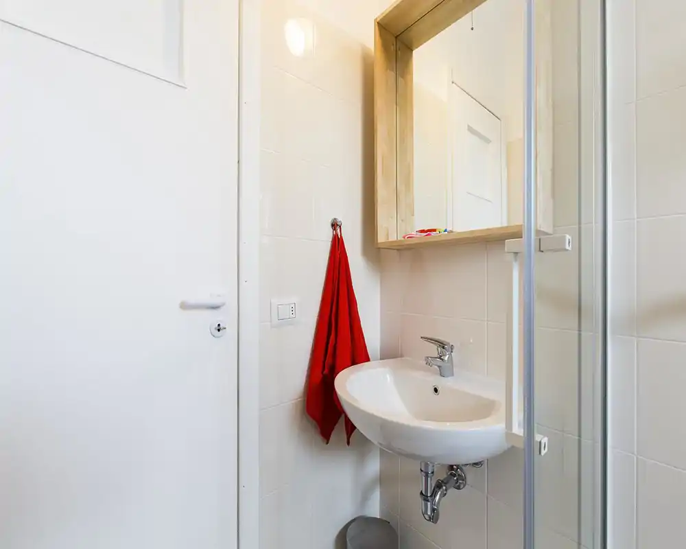 Bagno moderno compatto con lavabo sospeso, specchio con anta e parete doccia in vetro; piastrelle bianche e un asciugamano rosso a contrasto.