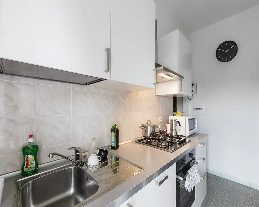 Cucina compatta e moderna con lavello in acciaio, piano cottura a gas, microonde e pensili bianchi — luminosa, funzionale e leggermente vissuta.