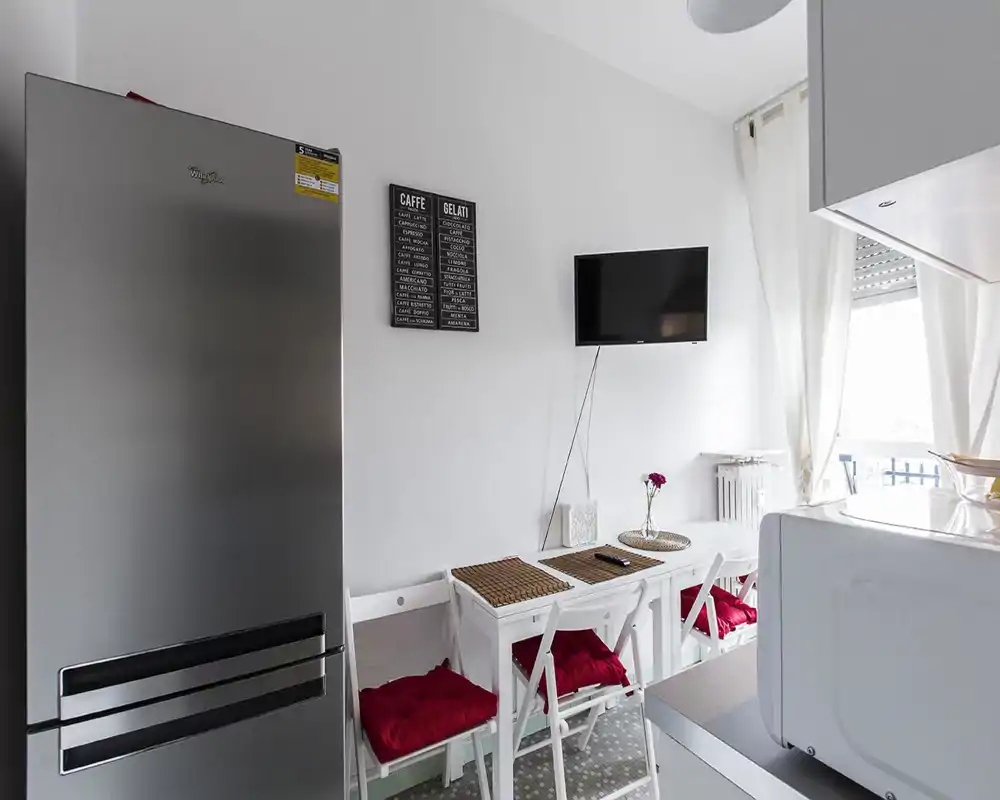 Angolo cucina moderno e compatto con frigorifero in acciaio, piccolo tavolo bianco con cuscini rossi, TV a parete e luce naturale dalla finestra/terrazzino.