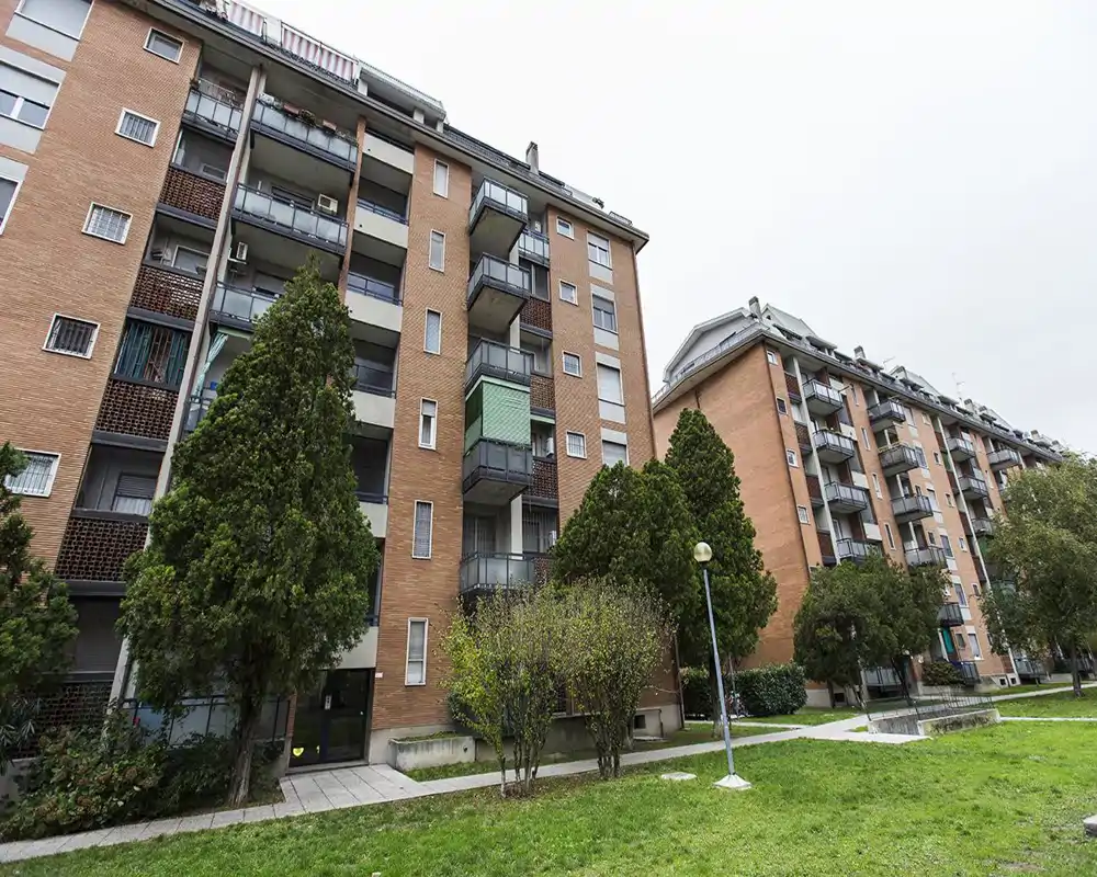 Vista esterna di un condominio in mattoni con balconi e un cortile comune erboso; alberi e un percorso pedonale conducono all'ingresso.