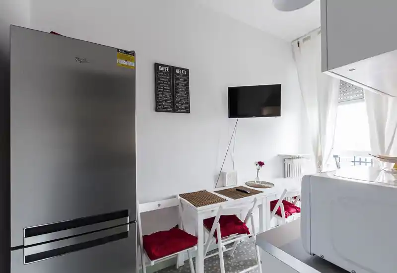 Angolo cucina moderno e compatto con frigorifero in acciaio, piccolo tavolo bianco con cuscini rossi, TV a parete e luce naturale dalla finestra/terrazzino.