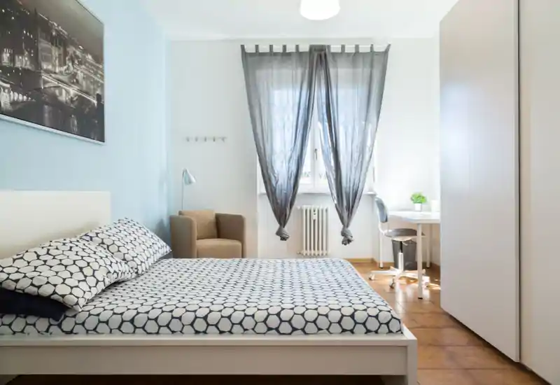 Bright, modern bedroom with patterned bedding, a cozy armchair, desk by the window and a large wardrobe — ideal for comfortable short or long stays.
