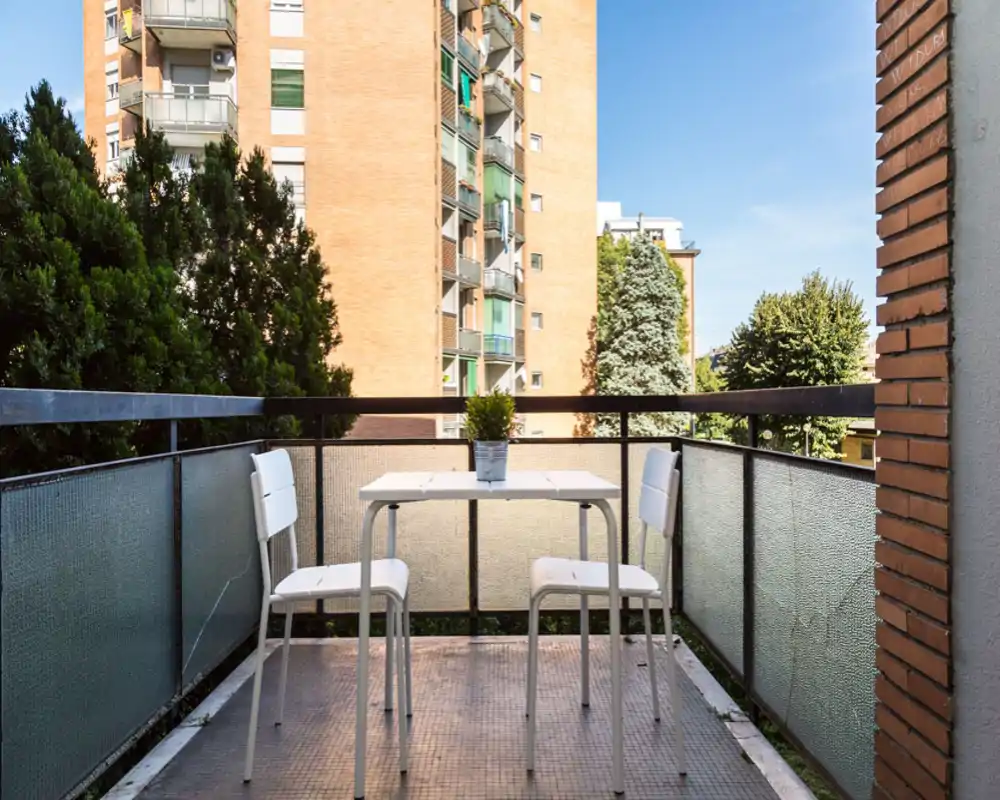 Sunny private balcony with a small table and two chairs, ideal for morning coffee or an evening drink. The view includes nearby residential buildings and mature trees, offering a quiet urban outlook.