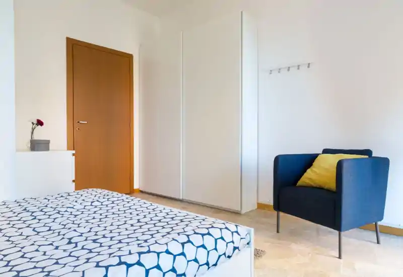 Bright, minimal bedroom with a patterned bedspread, white wardrobe and a navy armchair with a yellow cushion — clean, airy and simply styled for a relaxing stay.