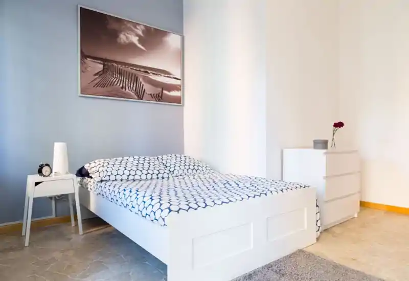 Bright, modern bedroom with a white platform bed dressed in patterned bedding, a small nightstand with lamp and alarm clock, and a three-drawer dresser with a single flower — clean, airy and well styled for rentals.