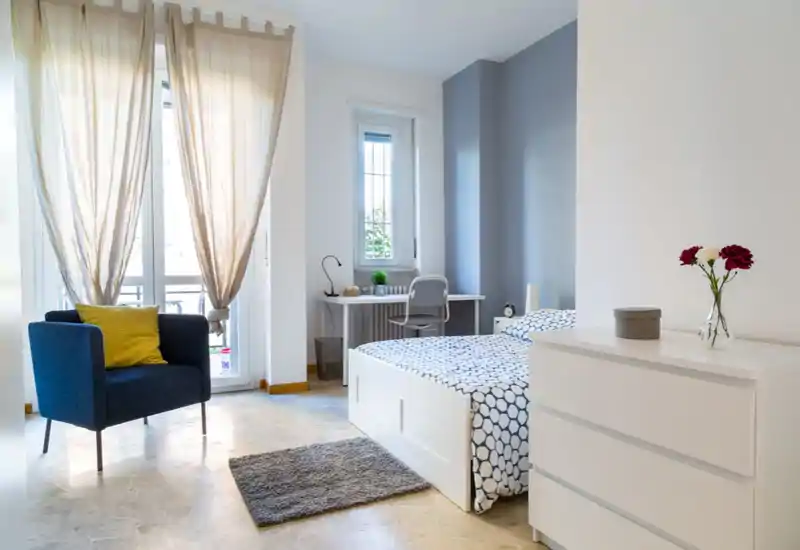 Bright, modern bedroom with a double bed dressed in patterned linens, a white chest of drawers, a cozy armchair by the window and a small desk — ideal for comfortable stays with natural light.