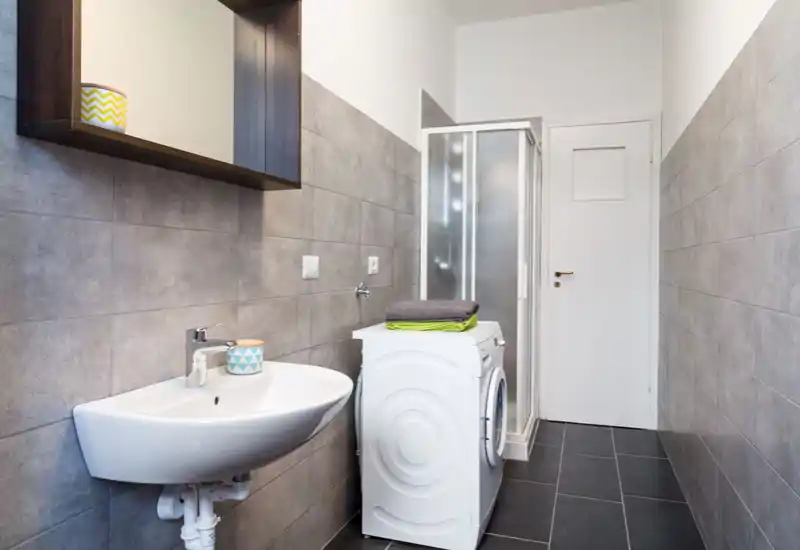 Modern tiled bathroom with a wall-mounted sink, shower cubicle and a washing machine; neutral grey tiles and neat, minimal styling.