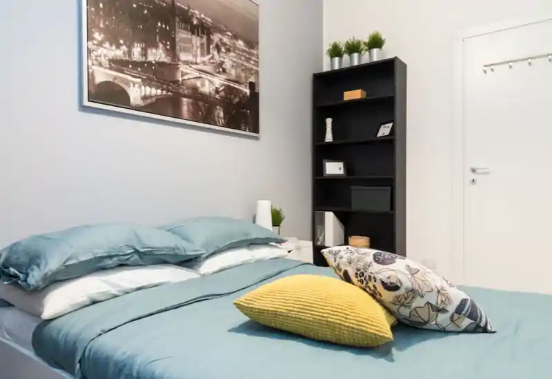 Camera da letto luminosa e ben allestita con un letto matrimoniale in lenzuola turchesi, cuscini decorativi e una libreria alta accanto a una porta bianca. L'ambiente risulta fresco e accogliente con un quadro a parete e piante discrete.