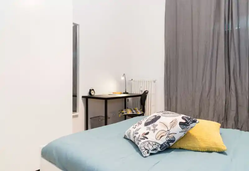 Camera da letto moderna e luminosa con letto ben rifatto, cuscini decorativi e una piccola scrivania lungo la parete — ideale per soggiorni confortevoli e smart working.