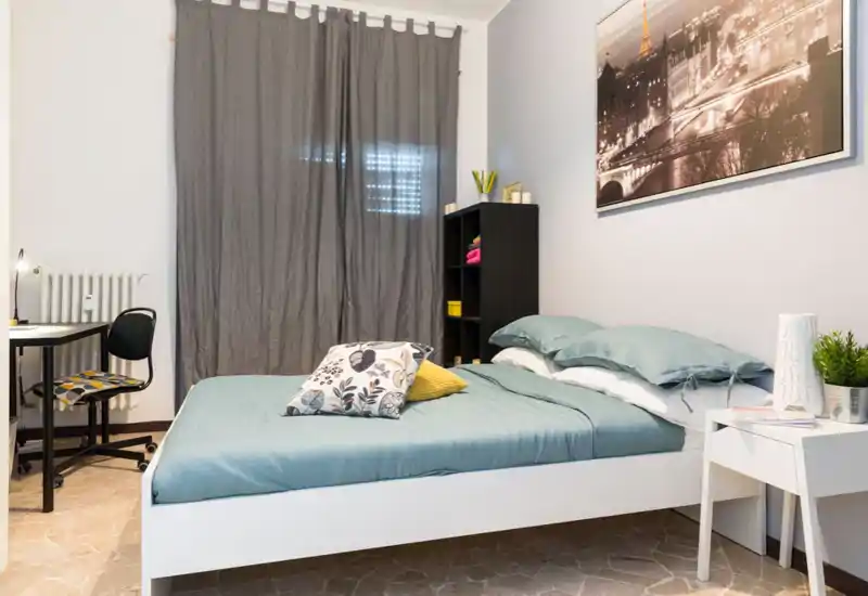 Camera da letto luminosa e ordinata con letto matrimoniale rifinito in tessuti verde acqua, cuscini decorativi, comodino con lampada e pianta e una scrivania angolare — ideale per riposo e lavoro leggero.