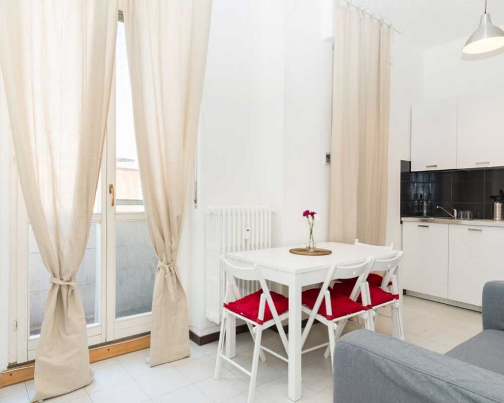 Angolo pranzo luminoso e moderno con tavolo bianco, sedie pieghevoli con cuscini rossi e un piccolo centrotavola floreale accanto alla cucina e alla porta del balcone.