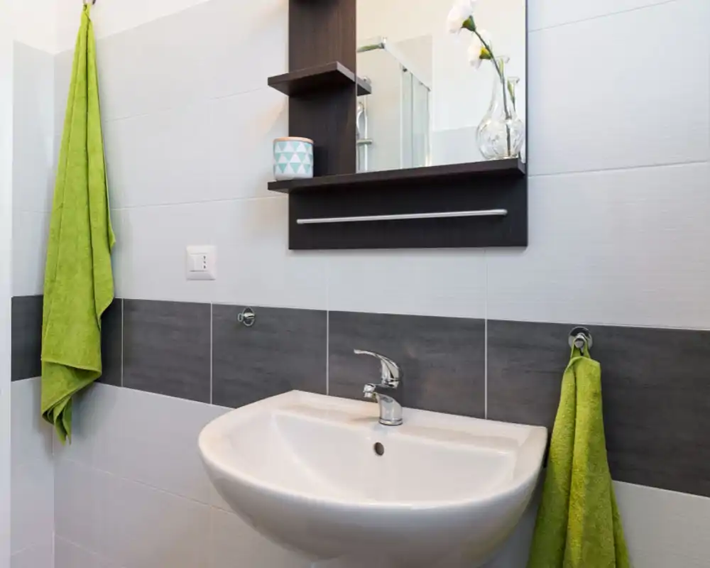 Area lavabo di un bagno moderno e luminoso con lavabo bianco, rubinetto cromato, specchio con mensole e asciugamani verdi come tocco di colore.