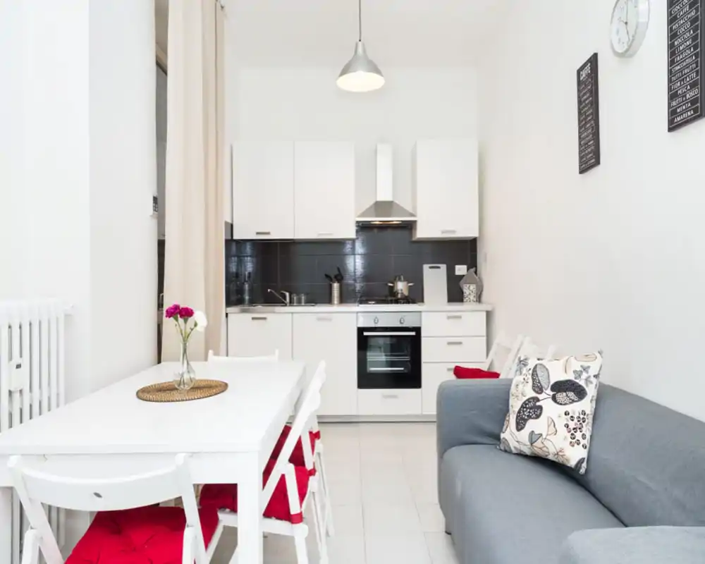 Zona giorno open space luminosa con angolo cottura bianco compatto, tavolo da pranzo e divano grigio accogliente con cuscini decorativi — moderna e ariosa.