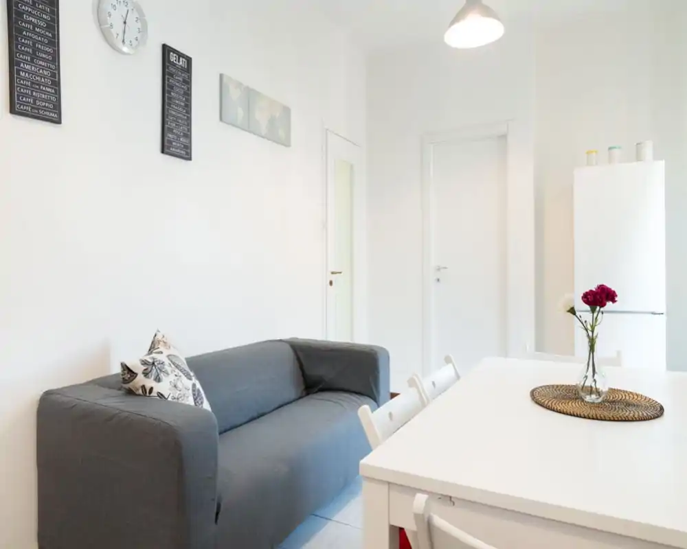 Zona giorno luminosa e minimal con divano grigio confortevole, cuscino decorativo e tavolo da pranzo in primo piano — perfetta per un appartamento compatto.