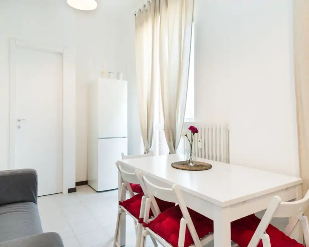 Zona pranzo luminosa e minimal con tavolo bianco, sedie pieghevoli con cuscini rossi e un vaso decorativo; la luce naturale che entra dalle tende crea un ambiente arioso e tranquillo.