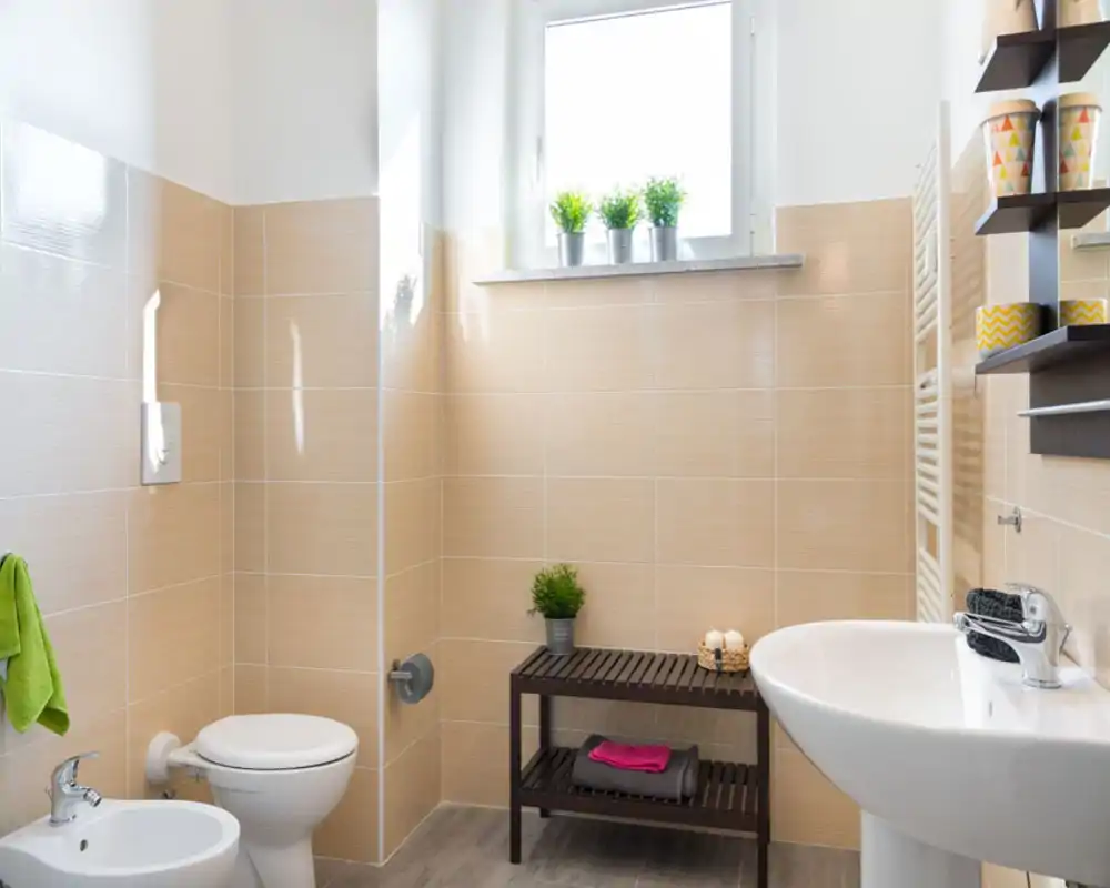 Bagno moderno e luminoso con piastrelle beige, wc sospeso, bidet e lavabo a colonna. Mensole e piante decorative donano un aspetto fresco e curato.