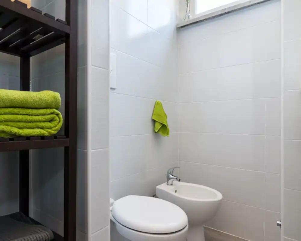 Piccolo bagno moderno con wc e bidet, pareti piastrellate bianche e scaffale con asciugamani verdi piegati.
