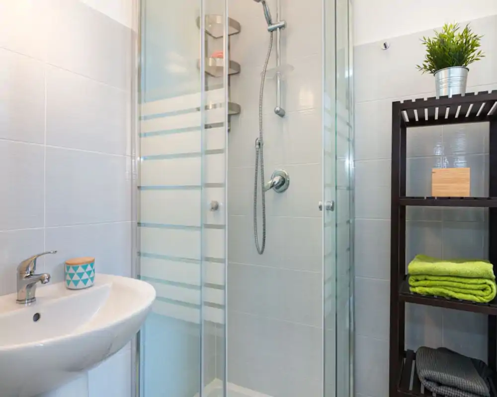 Bagno moderno e compatto con doccia a cabine in vetro, miscelatore a parete e piccolo lavabo; scaffale scuro con asciugamani piegati e una pianta per un tocco fresco.