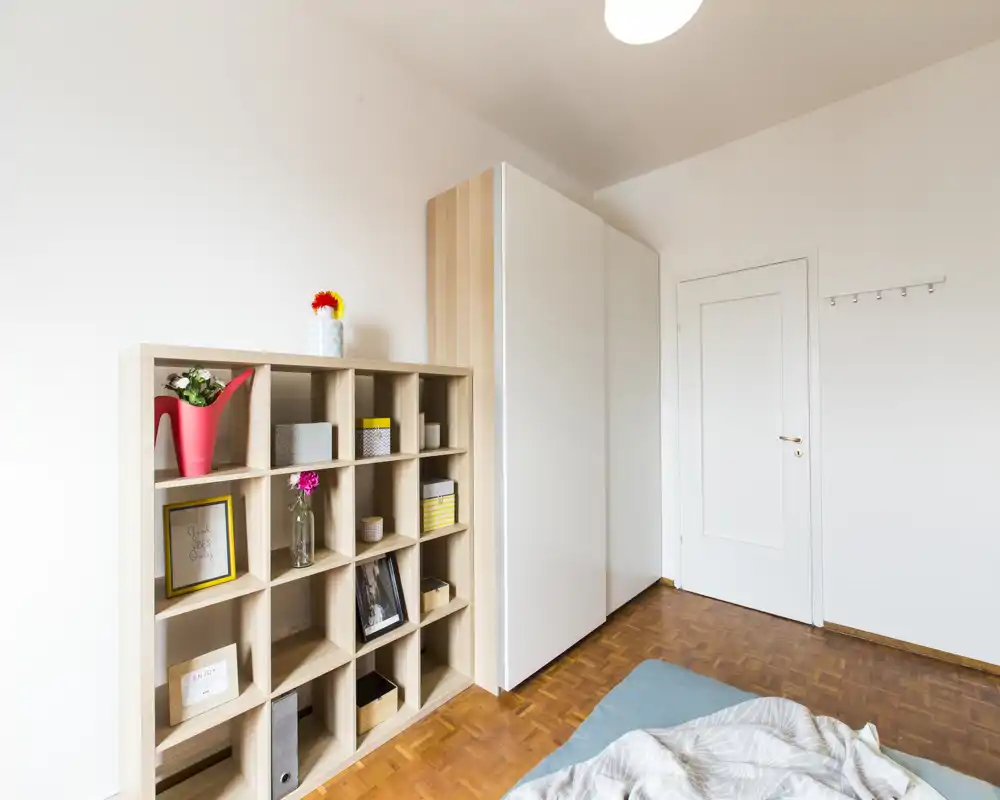 Bright, minimal bedroom corner with a visible bookshelf, wardrobe and the edge of a made bed; clean parquet flooring and soft natural light.