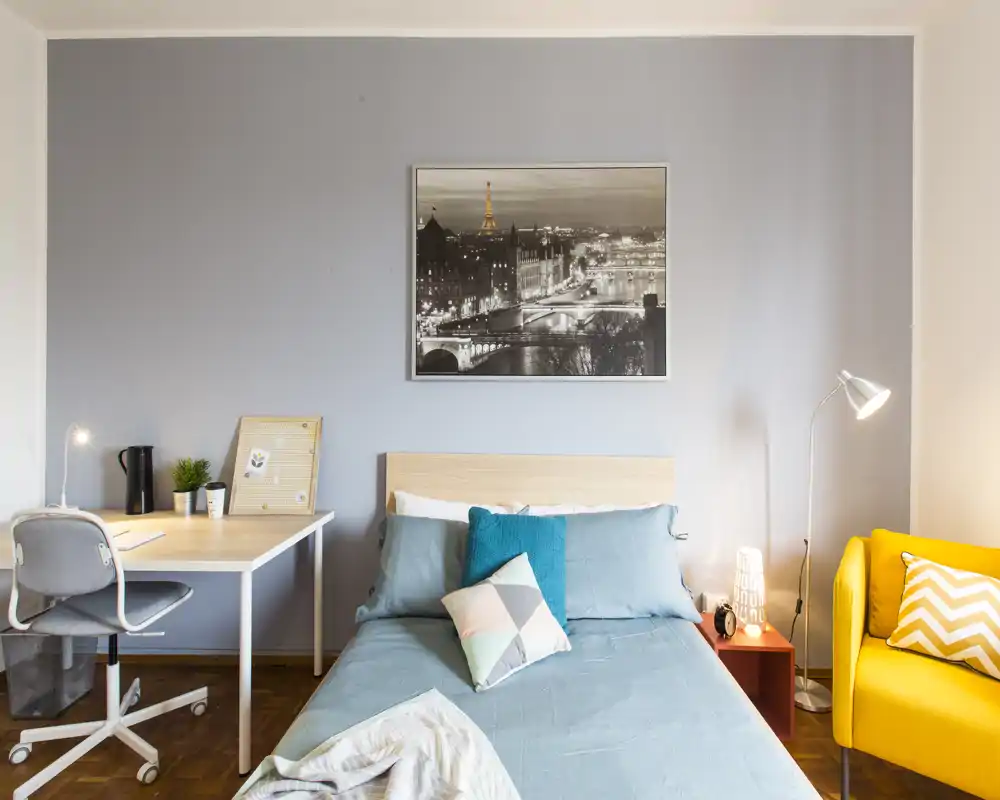 Bright, neatly staged bedroom with a double bed dressed in blue linens, decorative cushions and a cozy yellow armchair. A desk and lamp provide a practical workspace while a framed cityscape above the bed adds character.