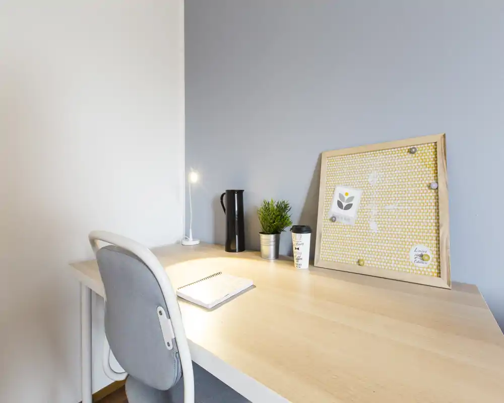 Bright, modern desk corner with a light wood tabletop, gray chair, small lamp, plant and a pinboard — ideal for a cozy workspace in a living area.