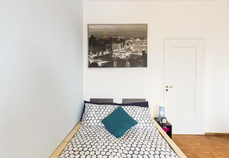 Dormitorio moderno y luminoso con cama doble con ropa de cama estampada, un cojín decorativo turquesa y mesita de noche con lámpara. Arte minimal en la pared y suelo de parquet crean un ambiente limpio y acogedor.