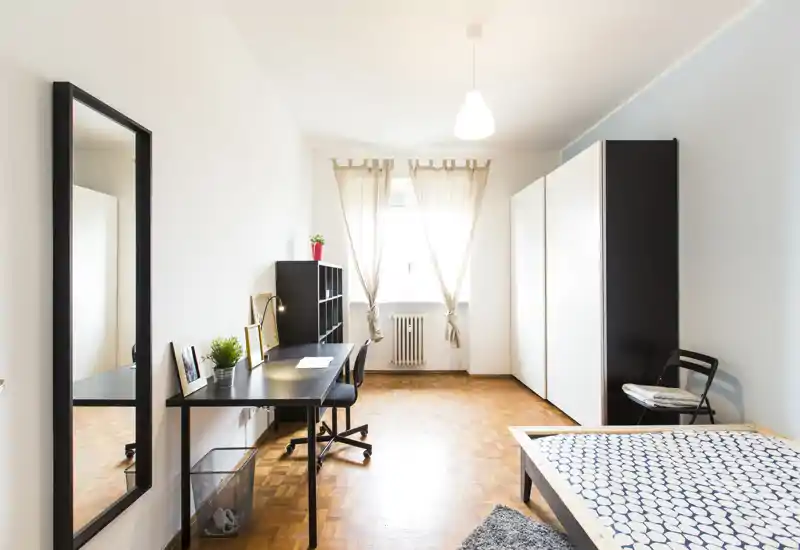 Dormitorio luminoso y minimalista con una cama individual en primer plano, escritorio y estantería junto a la ventana y un gran armario. La luz natural y los tonos neutros crean un espacio aireado y funcional, perfecto para estudiar y descansar.