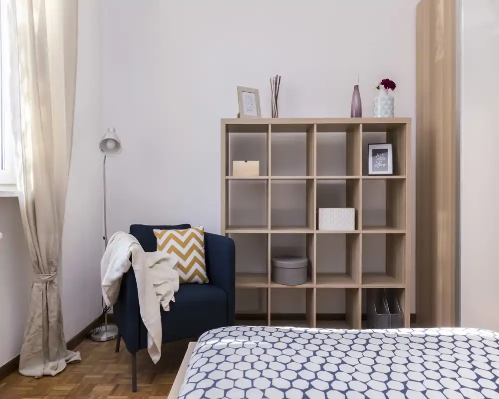 Angolo dormitorio luminoso e ben arredato con copriletto a motivo in primo piano, poltrona blu con cuscini e libreria a cubi in legno con oggetti decorativi.