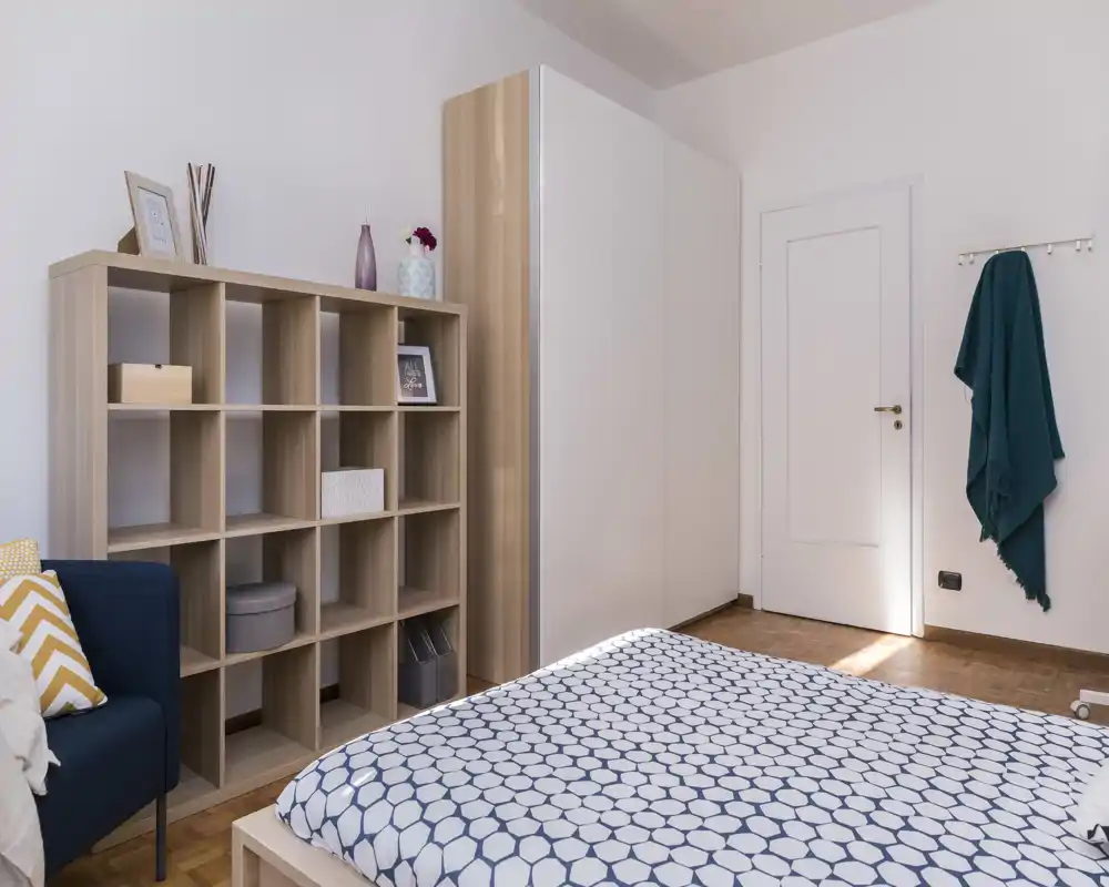 Camera da letto luminosa e ordinata con copriletto a fantasia, scaffalatura in legno e un grande armadio; linee pulite e luce naturale la rendono accogliente.
