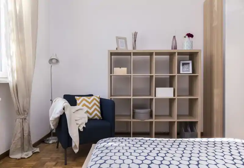 Bright, well-styled bedroom corner featuring a patterned bedspread in the foreground, a navy armchair with cushions and a wooden cubed shelving unit with decorative items.