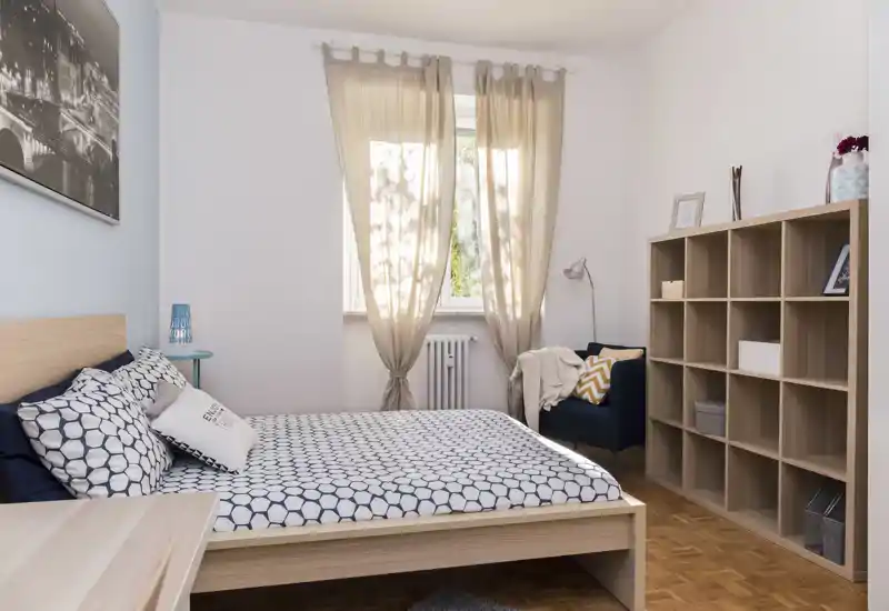 Bright, well-staged bedroom with a double bed, patterned bedding, a reading chair and open shelving; natural light from the window creates a welcoming atmosphere.