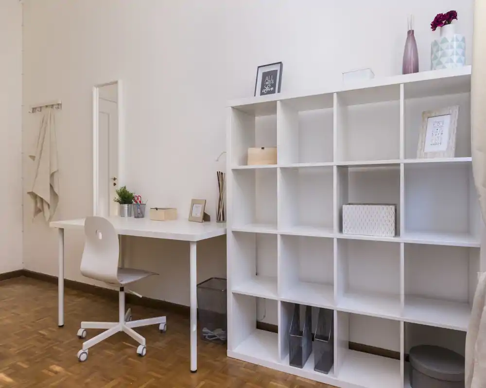Rincón luminoso y minimalista de sala/estudio con escritorio blanco, silla y estantería modular para almacenamiento, sobre suelo de parquet claro.