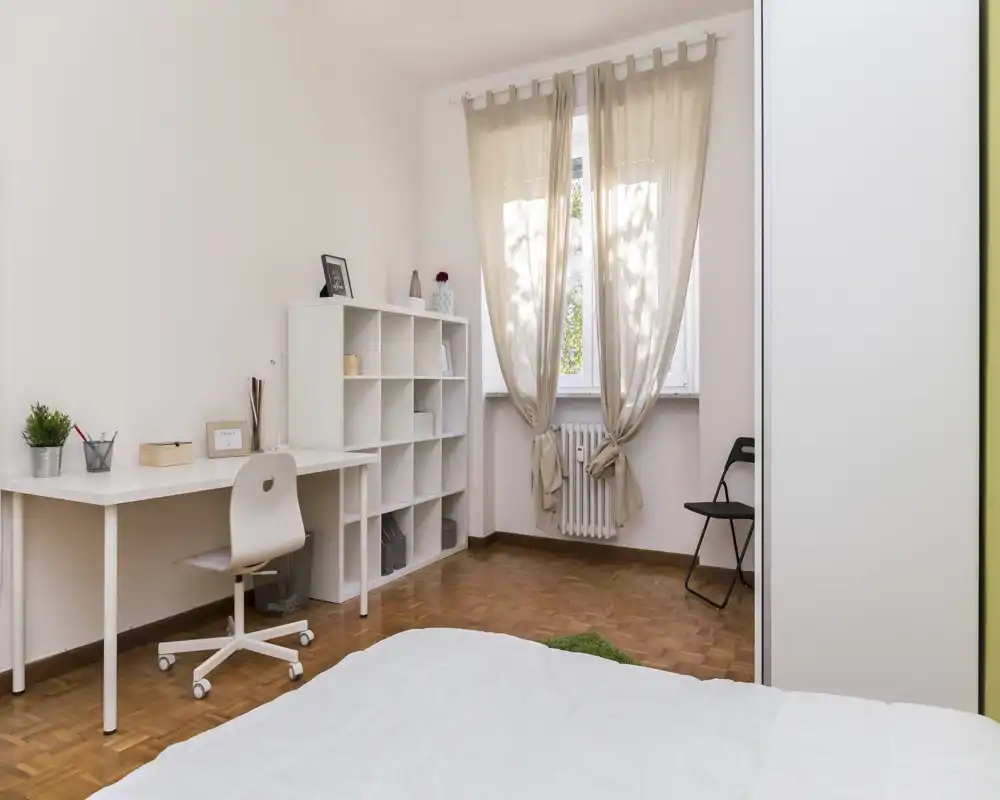Dormitorio luminoso y minimalista con la cama en primer plano, un escritorio blanco y estantería junto a la ventana que deja entrar luz natural suave.