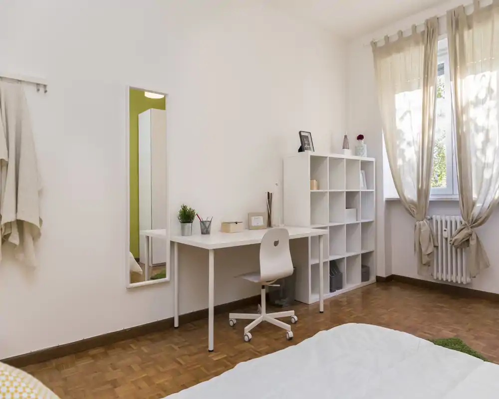 Rincón de dormitorio luminoso y ordenado con escritorio blanco, estantería modular y cama parcialmente visible en primer plano; las cortinas suaves y la luz natural generan un ambiente sereno y acogedor.