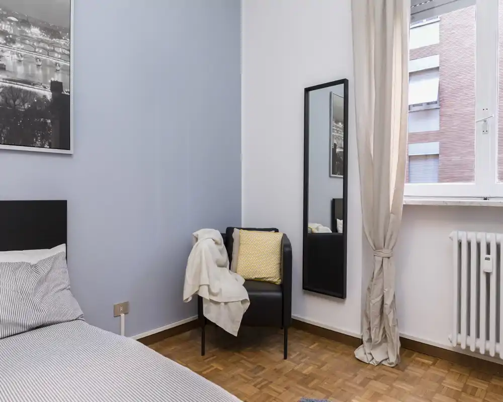 Angolo di camera luminoso e curato con letto comodo, poltrona con cuscino e coperta, specchio a figura intera e finestra che apporta luce naturale.