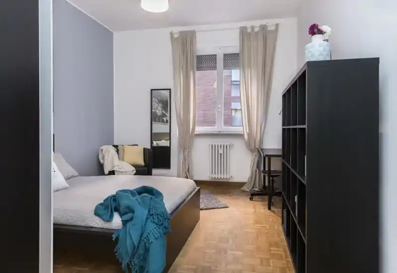 Camera da letto luminosa e ben arredata con letto matrimoniale, biancheria morbida e un plaid turchese; luce naturale dalla finestra e scaffalatura elegante rendono l'ambiente accogliente.