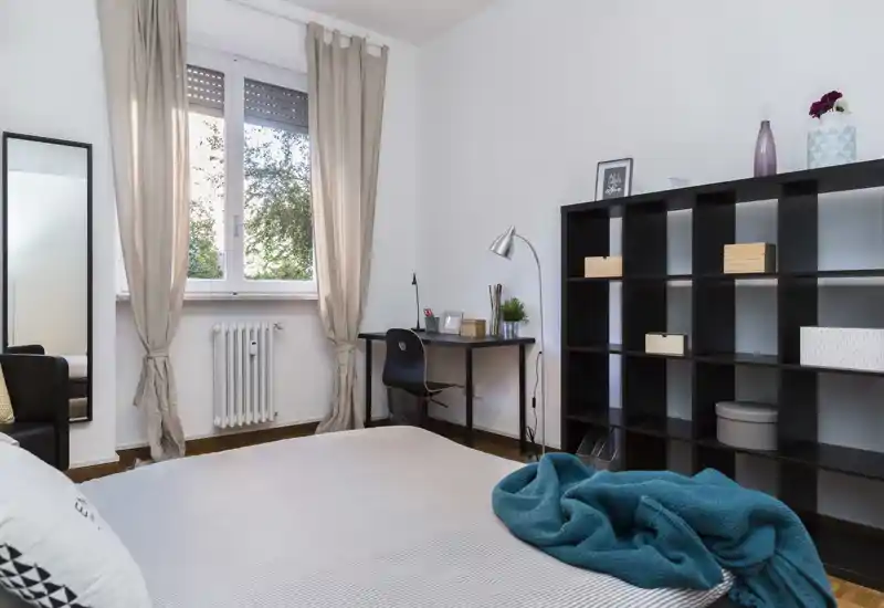 Camera da letto luminosa e ordinata con letto in primo piano, una scrivania vicino alla finestra e scaffalatura modulare per riporre oggetti.