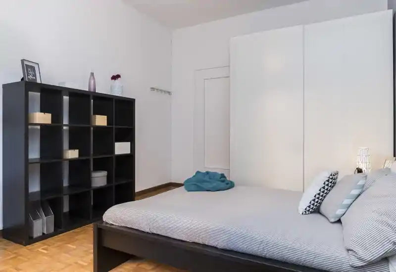 Camera da letto moderna e ordinata con letto matrimoniale, cuscini decorativi e un ampio armadio; colori neutri e una libreria modulare per contenere oggetti.