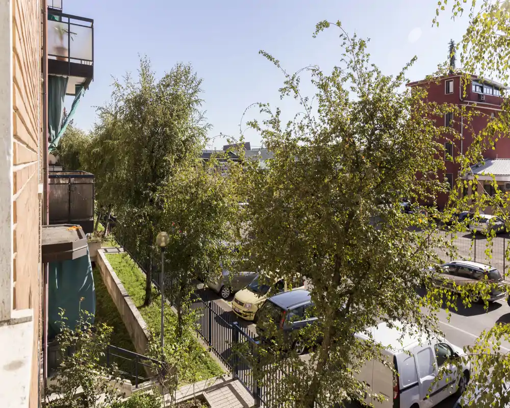 Vista soleggiata da un balcone su una strada fiancheggiata da alberi e auto parcheggiate, con uno scorcio urbano luminoso e edifici vicini.