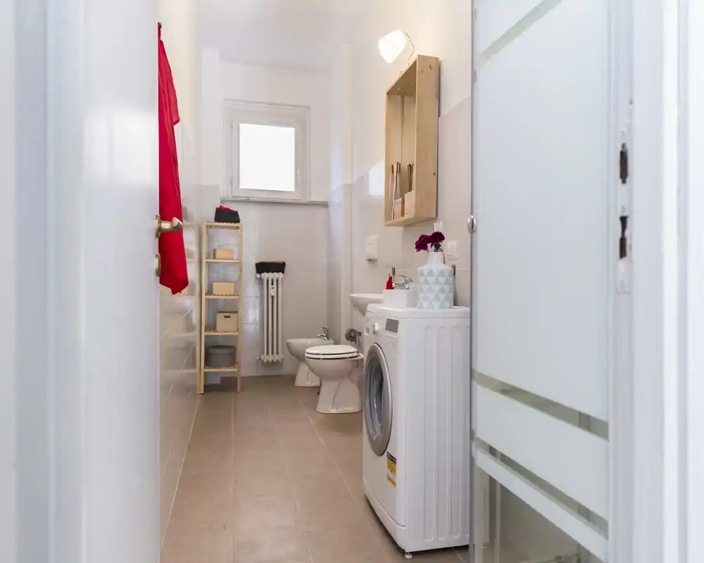 Bagno compatto e luminoso con lavatrice, WC e bidet; piastrelle neutre e mensole in legno donano un aspetto funzionale e pulito.