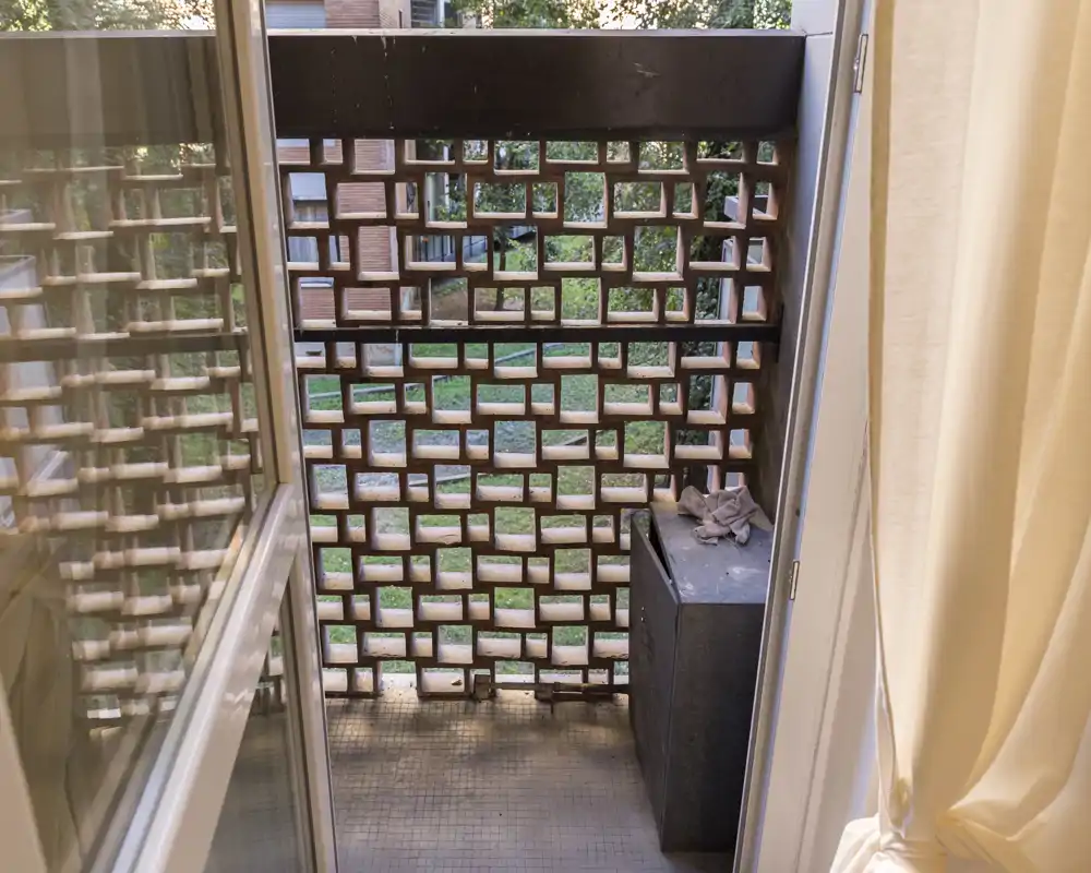 Piccolo balcone privato con griglia in mattone decorativa, pavimento piastrellato e un cassone per riporre oggetti; luce naturale e vista su un cortile verde.