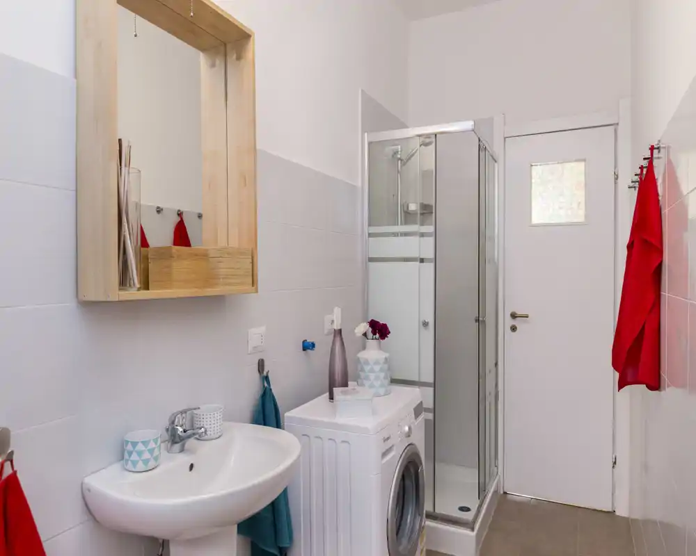 Bagno moderno compatto con cabina doccia angolare, lavabo e lavatrice a carica frontale; piastrelle chiare e asciugamani colorati donano un aspetto fresco e ordinato.