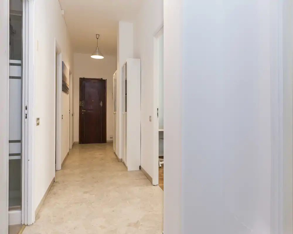 Corridoio d'ingresso dell'appartamento ben illuminato con pavimento piastrellato, lampada a sospensione e porta d'ingresso in legno.
